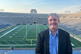 Photo of the author’s husband at the Notre Dame football stadium with her husband dressed in a suit and smiling.