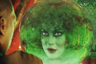 This is a screenshot from the movie the haunted mansion. It shows a woman’s head in a large glass orb. She is wearing bring red lipstick and dark eye liner. She has curly hair. She is looking at a man who is mostly out of the frame of the image.