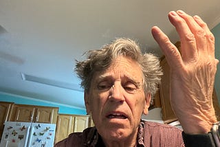 A man with very disheveled gray hair and a frustrated expression is sitting in a kitchen. He is raising one hand in an exasperated gesture, with his mouth slightly open. The background features wooden kitchen cabinets, a white refrigerator adorned with butterfly magnets, and a ceiling fan.