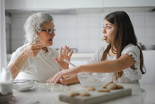 Woman with gray hair speaking to young girl with cookie in her mouth at kitchen table