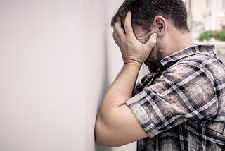 A frustrated man holds his head in his hands and leans against a wall.