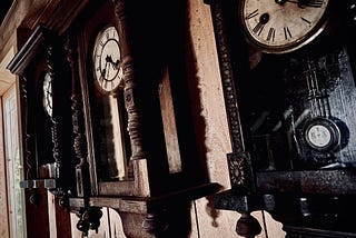 Three very old old clocks hanging on a wooden wall near a door with light coming in.