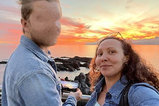 Beautiful colors of a sunset with the sky in the upper half of the photo and water in the lower half with the woman on the right smiling while looking into the camera and the man on the left with his face blurred out.