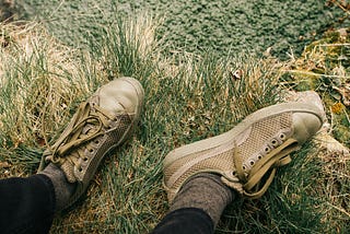 Top-down view of a person’s feet in worn, olive-green sneakers with loose laces, resting in patchy grass near a stone edge. The person’s legs are relaxed and slightly apart, as if pausing or observing from the sidelines.
