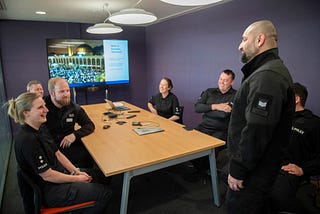 Six MDP officers, men and women, gathered in a conference room, sitting around a table, listening to PS Rahat who is standing and presenting about Ramadan. In the background there is a large digital screen with the image of a slide on it from PS Rahat’s presentation.