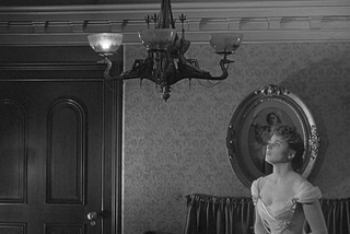 A black and white still from the 1944 film Gaslight. Paula, played by Ingrid Bergman, stares up with a haunted expression at a flickering gaslight chandelier in her home. Just one of its light is on.