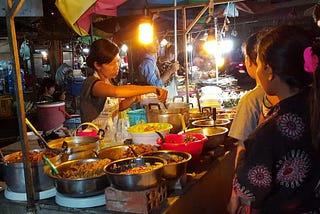 night market outdoor food stall in Siem Reap with woman serving from many bowls of food while customers look on