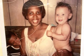 A Black woman smiling, looking straight into the camera while sitting on a couch with a pale skin baby in her left arm and a boy with a darker complexion sitting on her right, smiling and looking over at the baby.
