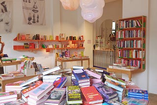 Interior of a bookstore. The tables are piled high with books by women and non-binary authors. In the background, a small kiosk serves coffee and cakes.