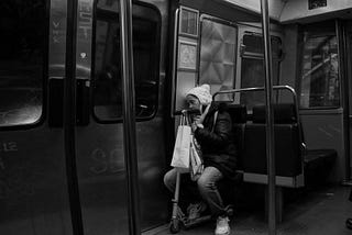 Foto preto e branco de mulher dormindo em um banco do metrô com um patinete e sacolas enquanto repousa sua cabeça na parede do vagão.