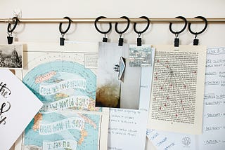 A collection of papers, maps, and images hangs from black clips on a gold curtain rod, creating a writer’s mood board. The papers include handwritten notes on a world map, a page of typed text with annotations, and a whiteboard with notes on story structure.