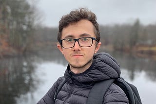 A man with black hair and glasses. He’s wearing a black jacket and standing next to a lake surrounded by trees. The sky is grey and he has a stoic facial expression.