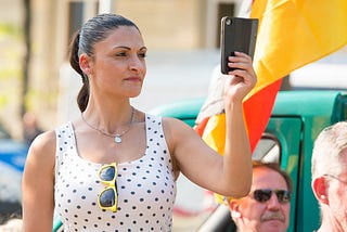 A Middle-Aged Woman With A Ponytail Wearing A Polka Dot Top Taking A Selfie In A Public Space.