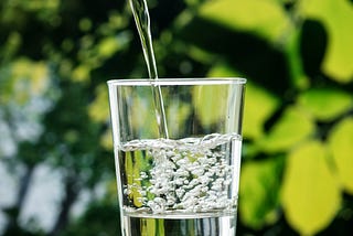 Close-up of water being poured into a clear glass, with bubbles forming as the glass fills. Green, blurred leaves are visible in the background, suggesting an outdoor setting.