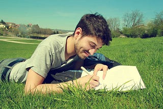 Man laying front down, on grass, writing with pen and paper