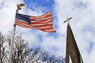 A U.S. flag hangs on an eagle-topped flagpole against a blue sky. To its right is a church steeple, nearly equal in height.