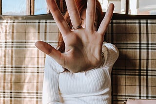 Woman with long read hair sitting on a plaid covered sofa. She is wearing a white ribbed sweater. Her left arm is out-strecthed and her hand is raised with palm facing the reader in the center of the photo.