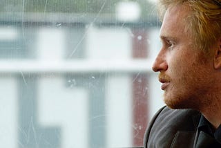 A close-up of a guy sitting on a bus, staring out the window. The view from the window is blurred, and the window has scratch marks.