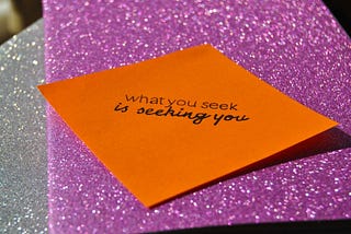 A sticky note with the inscription ‘What you seek is seeking you’ on a pink glitter background.