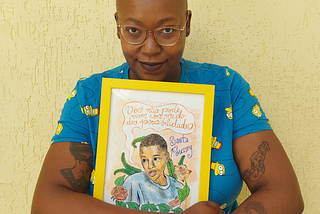 Na foto, uma mulher negra, careca, com uma camiseta azul, segura uma quadro escrito: você não pode ter medo das possibilidades