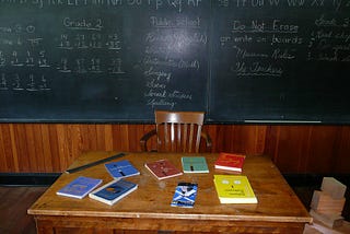 Image of books spread across a teacher’s desk in front of a classroom chalkboard.