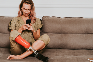 Person with orange prosthetic leg sitting on brown couch, legs crossed. Looking at a black cell phone in hands.