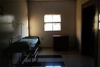 A bleak looking hospital room, with an empty bed and IV stand and a small window .