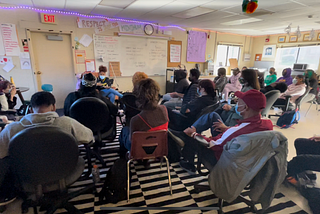 A view from behind a class of teenagers and one elder gathered into the shape of an audience. A student wearing a mask sits facing the rest.