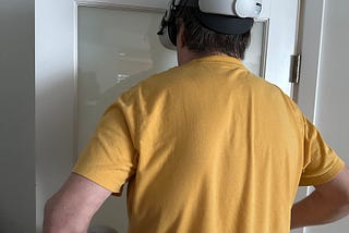 A tall man in a mustard-colored shirt walks into a door with a VR headset on.