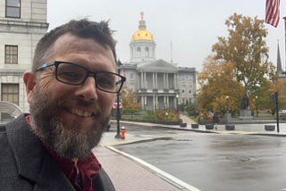 Author in a selfie across a rainy street from the NH Statehouse