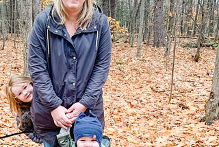 White, blond woman with glasses stands in a forest in autumn with two young children