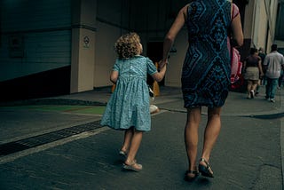 A girl, perhaps five years old, with blonde, curly hair and wearing a blue dress, is walking down a street. She is holding the hand of a woman in a blue dress.