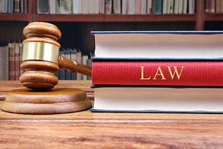 Wooden judge’s gavel resting on its sound block beside a stack of hardcover law books — one with a prominent red spine labeled “LAW” — on a polished wooden desk, framed by a blurred background of law-library shelves.