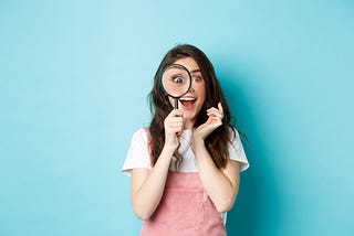 Happy young woman looking through a large magnifying glass with an excited face as if she has just discovered something. She is standing up, wearing pink dungarees and a white T-shirt against a blue background.
