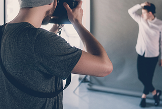 A photographer in a studio captures a male model posing against a gray backdrop during a professional photoshoot.