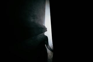 A woman’s silhouette stands behind a partially open curtain, bathed in soft light and mist.