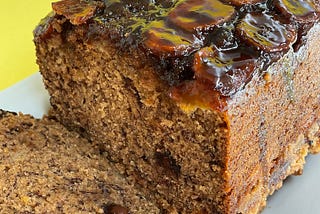 upside down banana bread with a slice cut, on a white plate sitting on a textured bright yellow background