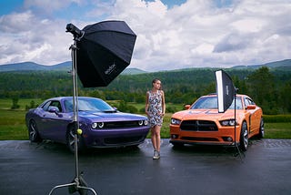 Behind the scenes of a fashion photoshoot showing flash equipment, model, and lots of detail in a Vermont summer scene with two modern muscle cars.