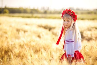 A girl in traditional Ukrainian attire stands in a wheat field. The poem featured in this article focuses heavily on the landscapes of Ukraine, particularly fields. The poem also focuses on a young girl with red ribbons in her hair and her journey to protect herself from outside forces. | Photo courtesy: Fokus_nik, Goodfon.