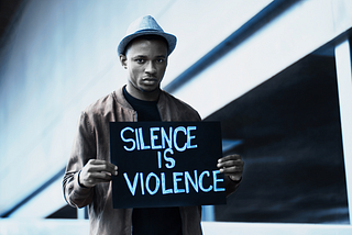 A Black man wearing a white hat and a brown suede bomber jacket holds a hand-written sign that says “Silence is Violence.”