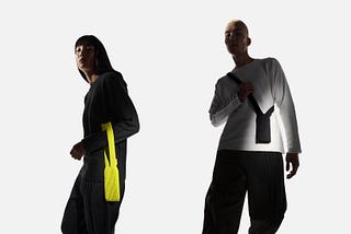 Two models pose against a plain light-gray background wearing modern, minimalist outfits. The person on the left wears a black long-sleeve top with a bright yellow fabric strap detail hanging from the side and loose black pants. The person on the right wears a white long-sleeve top with a black strap accessory draped across the chest and similar dark loose pants. The lighting creates strong shadows, emphasizing the clean, sculptural shapes of the clothing. (Apple photo)