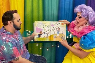 Two Drag Queen Story Time presenters are holding up a children’s book in front of a rainbow background as they read the story aloud
