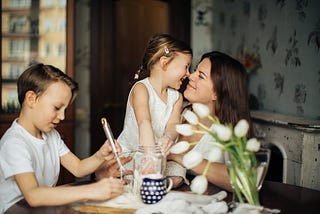 A mother with dark brown hair smiles as she hugs her brown-haired daughter. Her brown-haired son sits beside them at a table. There are white flowers on the table.