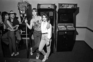 A young couple stand with their arms around one another in front of a pair of upright video games, while another couple lean against a pinball machine