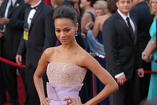 Actress Zoe Saldana in a sleeveless shimmering pink and lavender gown in front of a red rope.