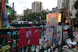 Image is a photograph of a subway station entrance/exit in downtown Manhattan by City Hall covered in hand painted signs about police brutality and abolishing the police. Text on signs includes: “Oink! Oink! Oink! Cops are Killers”, “Abolish the Police”, “Black Lives Matter”, and “Justice for Layleen Polanco”. Further in the distance beyond the subway entrance are a crowd of people gathered. Beyond the people are high rise buildings.