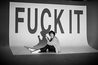 A black and white image of a man with curly hair, sitting on the floor with his legs stretched out, and looking at the camera. The projector projects on the man the words ‘FUCK IT’ on a white background.