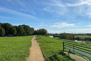 A gravel footpath stretching ahead between two grassy banks. It runs alongside the River Parrett, which is to the right of the picture. There are riverside benches and trees in the distance. It’s a sunny day with blue sky and wispy white clouds. This was taken at the very end of my half marathon run.