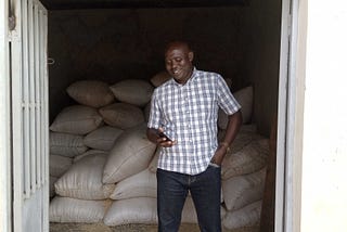 A African man looks down at the smartphone in his hand at the entrance of a storefront full of bags of grain