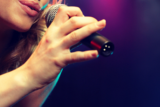 Singer sings into microphone held by their hand. Only their mouth is visible. Dark background with purple hue.